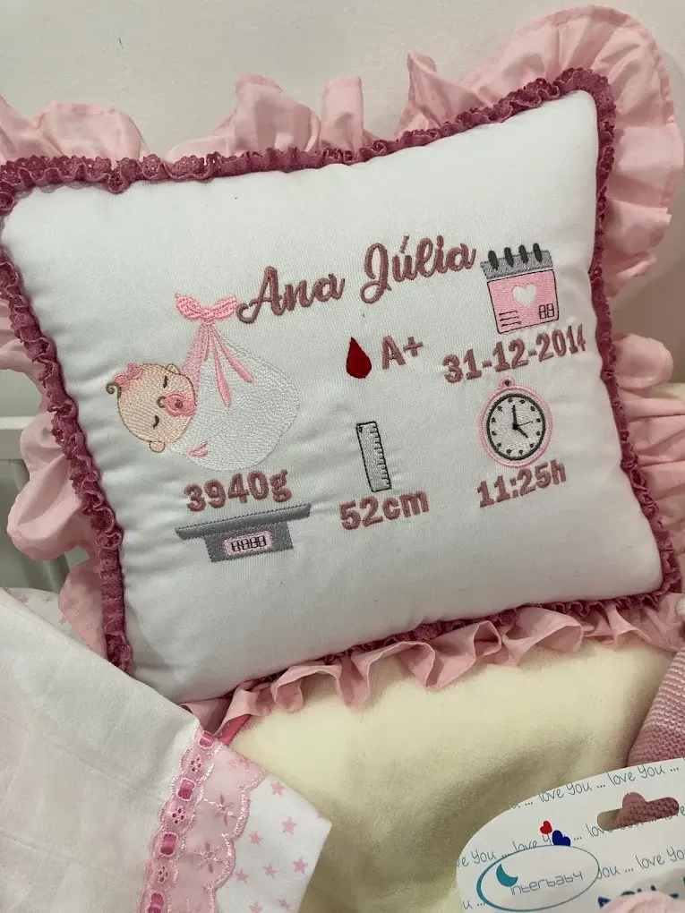 Almofada branca com folhos rosa e o nome Ana Júlia, data, hora, peso e medida de nascimento bordados.