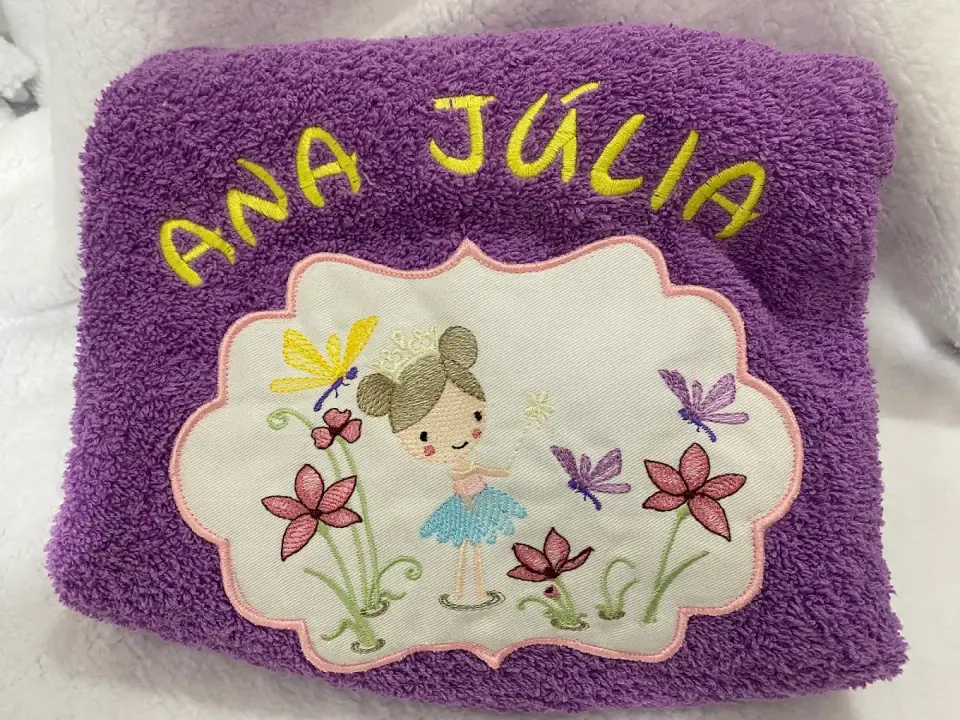 Toalha de felpo lilás com o nome Ana Júlia bordado a amarelo e uma bailarina entre flores.